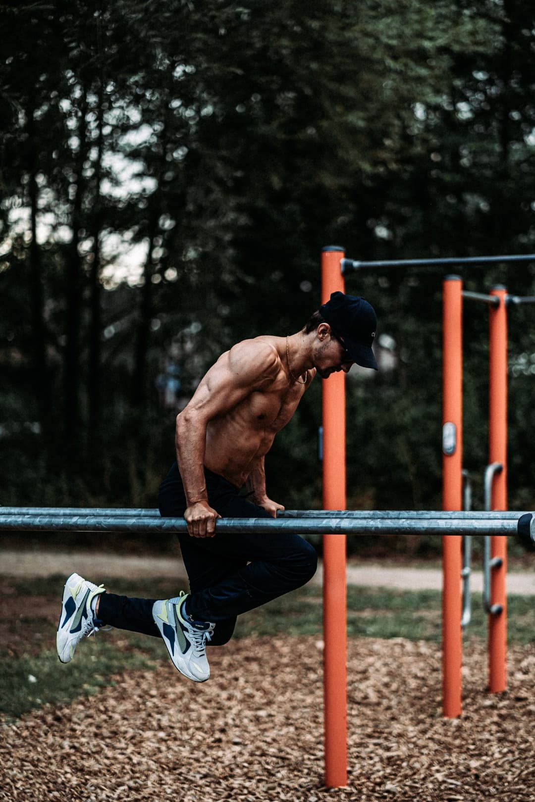 Shirtless man in a cap leaning far forward on low parallel bars at a wooded outdoor park, feet off the ground