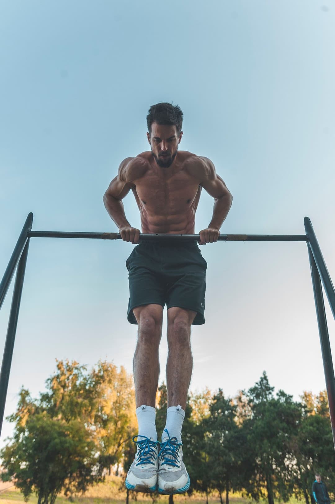 A shirtless man pressing up over a thin outdoor straight bar with bent arms, seen from below against a pale sky and trees