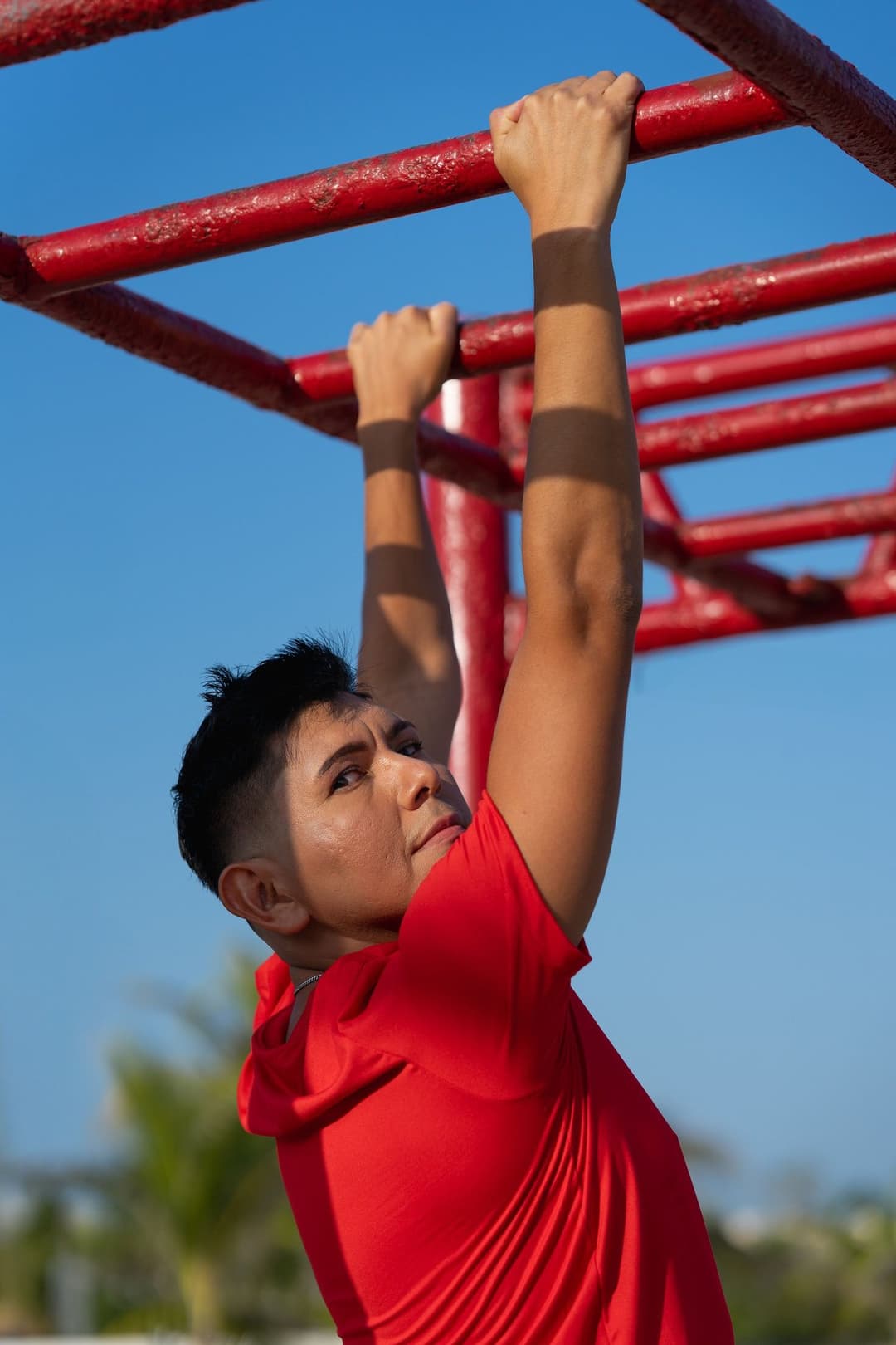A man in a red hoodie hanging from red monkey bars with an overhand grip and looking up at the bar