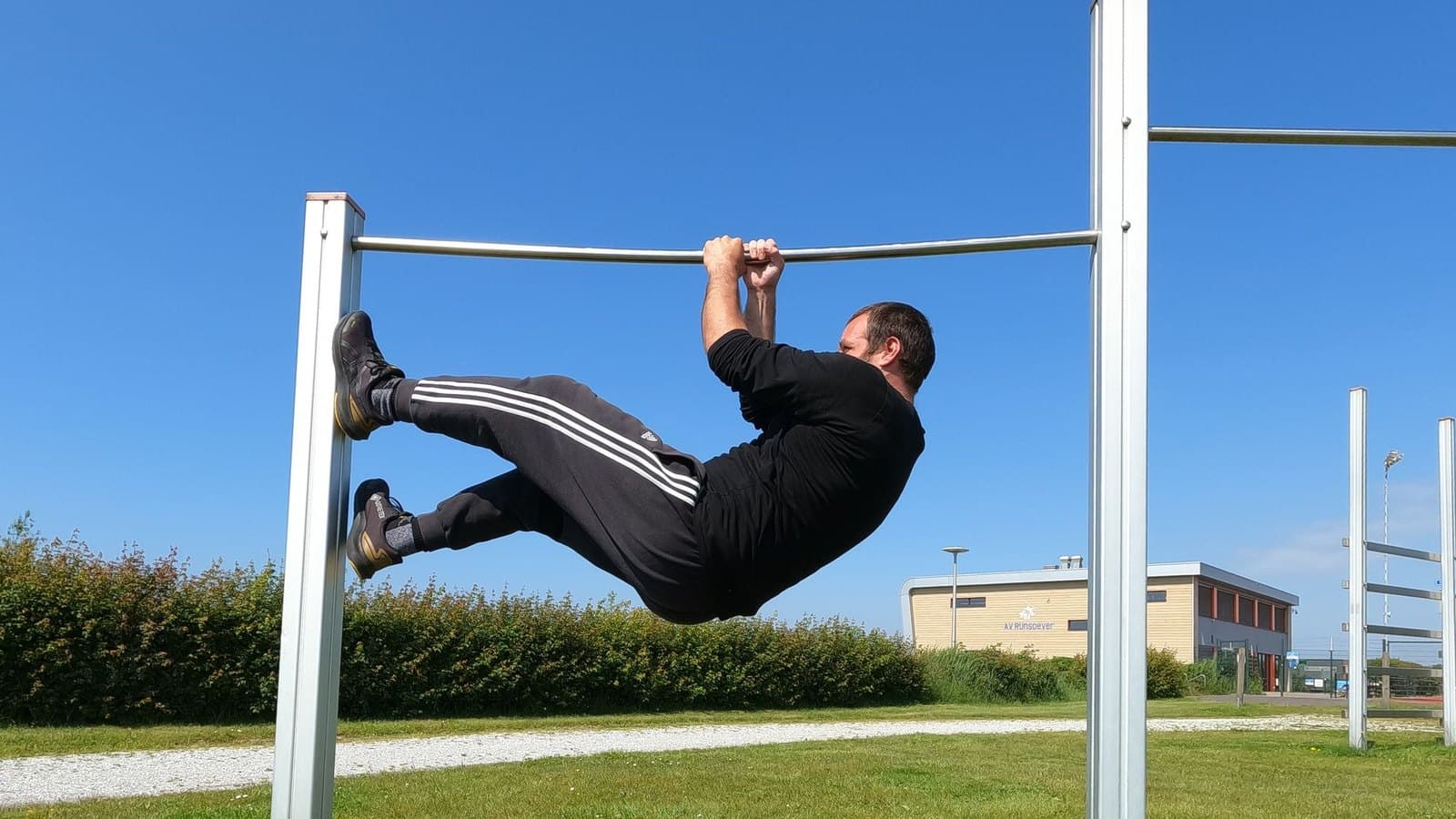 Man in a black tracksuit pulling his body close to horizontal under an outdoor pull-up bar on a sunny day