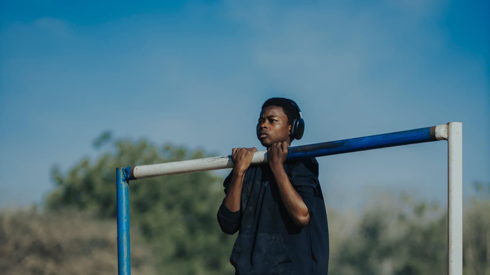 A young man in a black hoodie and headphones holding his chin above a blue and white pull-up bar in a dry field under a blue sky